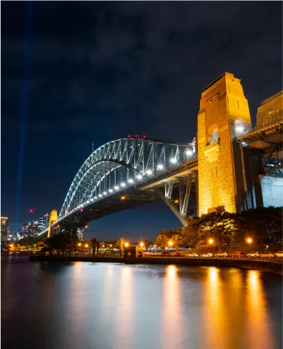 A lit-up arch bridge spans over water at night, with a large illuminated tower on the right and city lights in the background.