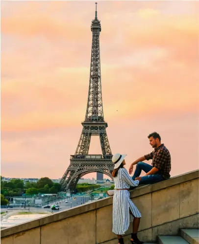 Two people sit on a ledge facing each other with the Eiffel Tower in the background at sunset. The woman wears a striped dress and hat; the man wears a plaid shirt and jeans.