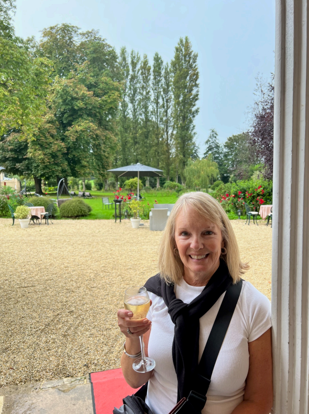 A woman stands smiling, holding a glass in an outdoor setting with a gravel area, greenery, tables, and trees in the background. She wears a white shirt and a black sweater draped over her shoulders.