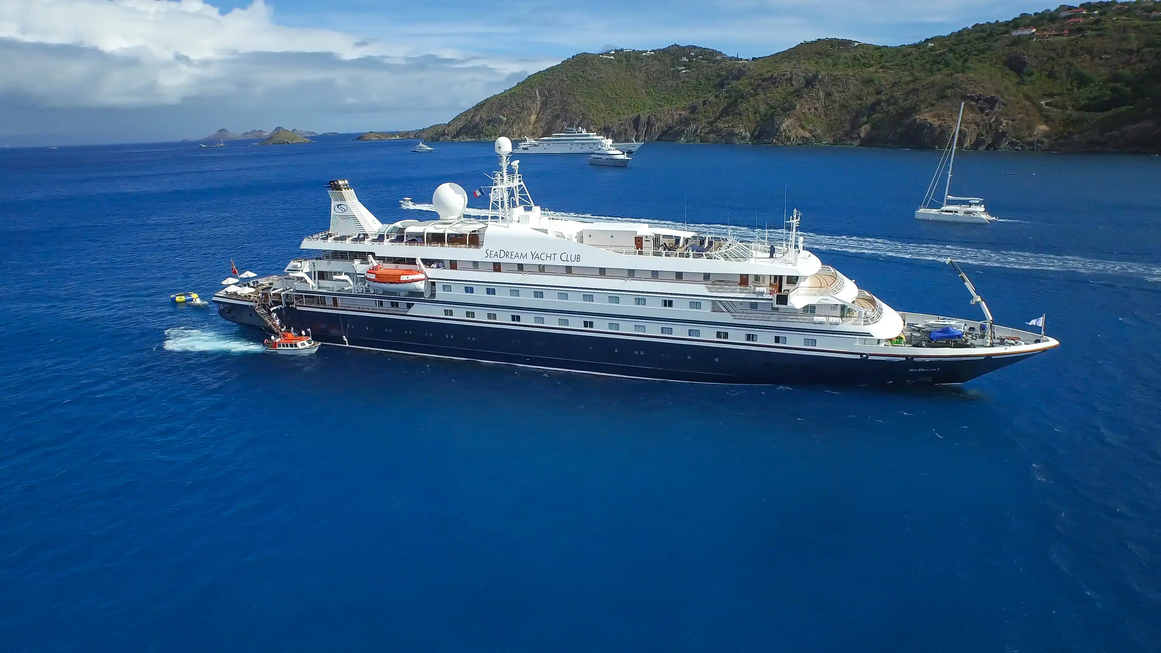 A large luxury yacht with white and navy details sails on a calm blue sea near green, hilly islands. Smaller boats are nearby.