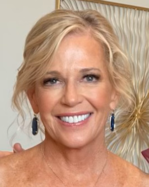 Smiling woman with short blonde hair, wearing blue earrings, in front of a patterned background.