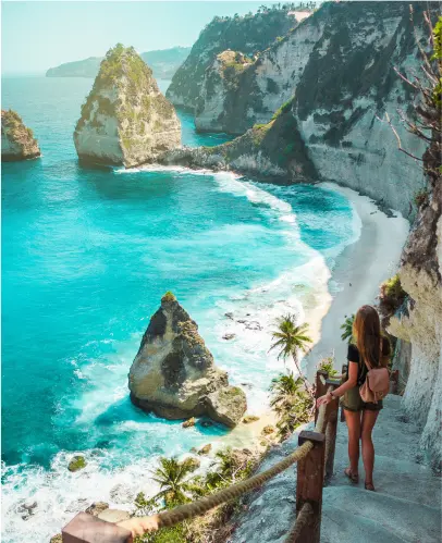 A woman stands on a cliffside path overlooking a turquoise ocean with rocky formations, white sandy beach, and palm trees on the shoreline.