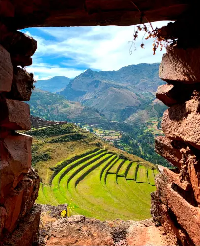 View through a stone window of terraced green fields on a hillside with mountains in the background under a blue sky.