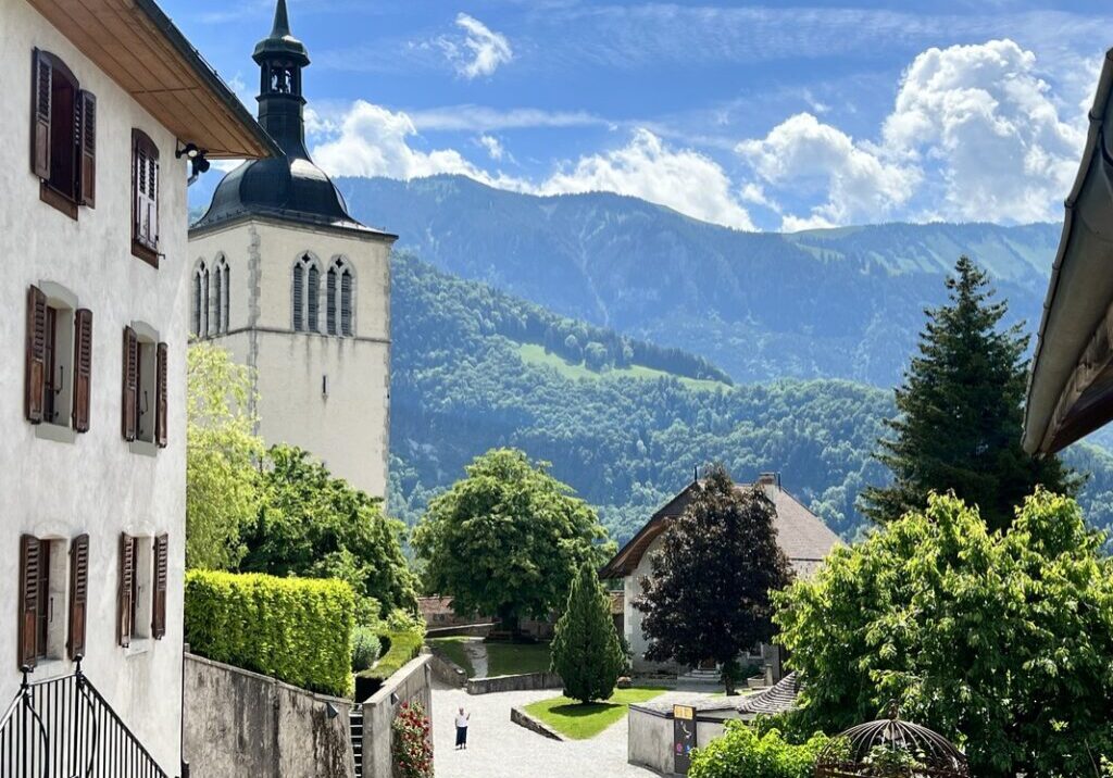 A scenic view of a village with a white church tower, traditional houses, lush greenery, and mountains in the background under a partly cloudy sky.
